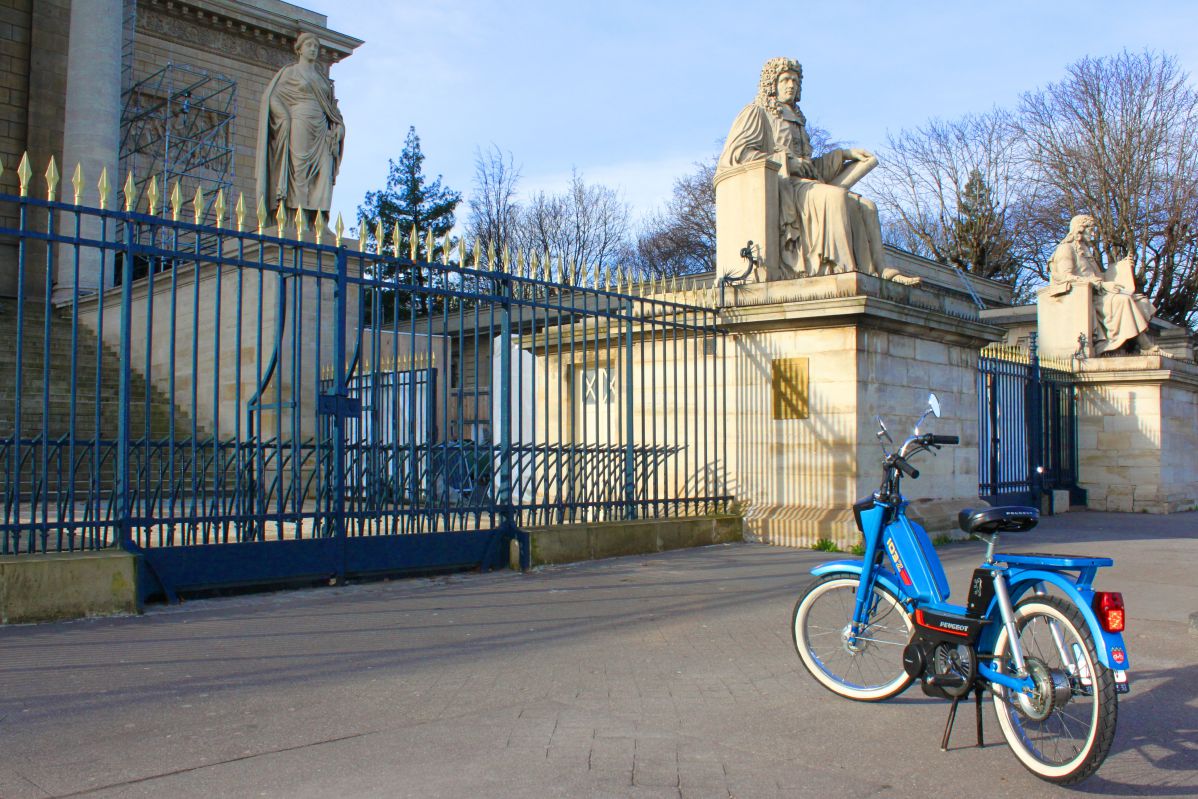 Une mobylette bleue devant la statue d'Henri François d’Aguesseau et l'Assemblée nationale, à Paris.