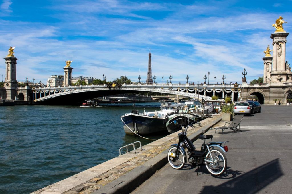 Mobylette sur un quai de Seine devant un péniche et le pont Alexandre 3.