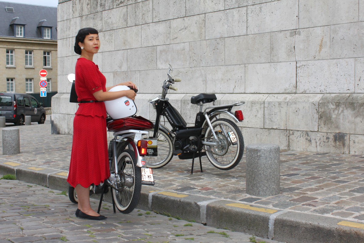 Une femme avec une robe rouge vintage devant des mobylettes à Paris